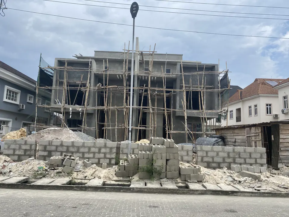 Two-storey house under construction behind a blockwork boundary wall, with timber scaffolding